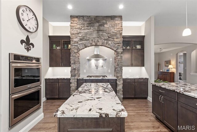 408 Hamlet Ct, Lake St Louis, MO 63367 - kitchen with stainless steel appliances, wall chimney range hood, dark brown cabinetry, light wood-style floors, and a kitchen island