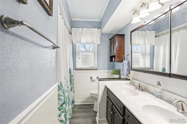 full bath with crown molding, toilet, double vanity, a wainscoted wall, and wood finished floors