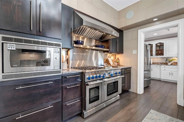 kitchen with appliances with stainless steel finishes, wall chimney range hood, light stone countertops, white cabinets, and tile walls