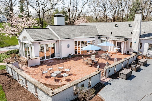 rear view of house featuring a chimney, a shingled roof, outdoor dining area, a patio, and brick siding