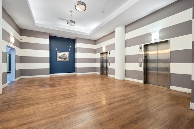 empty room with elevator, a raised ceiling, wood finished floors, and baseboards
