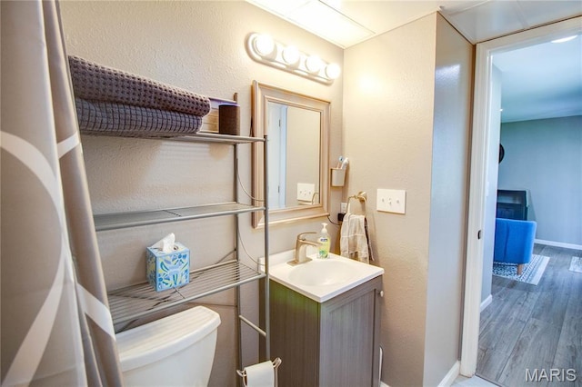 half bath featuring wood finished floors, toilet, vanity, baseboards, and a textured wall