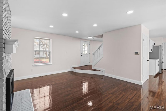 unfurnished living room with a brick fireplace, dark wood-style flooring, baseboards, stairway, and recessed lighting