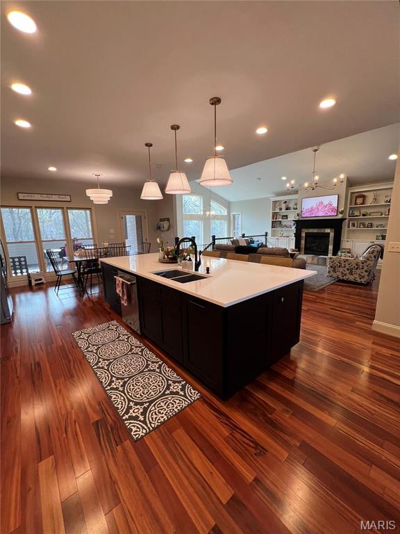 kitchen featuring a sink, dark wood-type flooring, a glass covered fireplace, an island with sink, and dishwasher