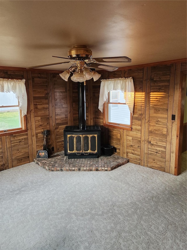 unfurnished living room featuring a wood stove, wood walls, plenty of natural light, carpet flooring, and ceiling fan