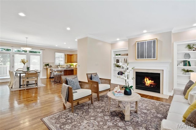 living room with crown molding, a fireplace with flush hearth, light wood-style flooring, built in features, and recessed lighting