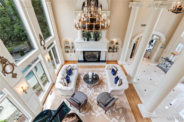 living room with a towering ceiling, a chandelier, ornate columns, wood finished floors, and a fireplace with raised hearth