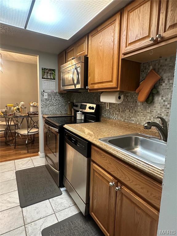 kitchen featuring stainless steel appliances, a sink, backsplash, light tile patterned floors, and brown cabinetry