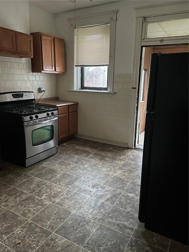 kitchen featuring stainless steel gas stove, freestanding refrigerator, brown cabinetry, tile walls, and wainscoting