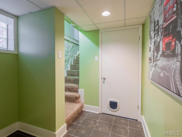hallway with a paneled ceiling, baseboards, tile patterned flooring, and stairway