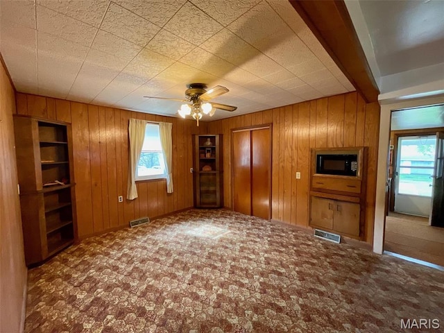 unfurnished living room featuring healthy amount of natural light, wood walls, a ceiling fan, carpet, and baseboards