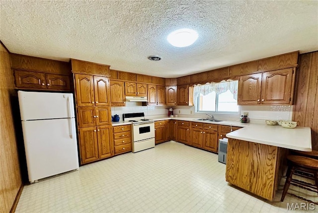 kitchen with white appliances, under cabinet range hood, brown cabinets, a peninsula, and a textured ceiling