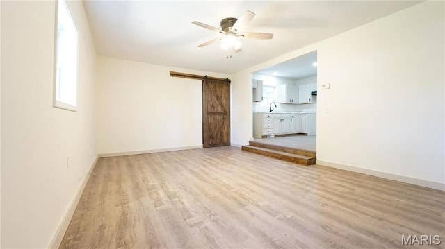 unfurnished living room with a barn door, light wood-style flooring, a sink, a ceiling fan, and baseboards