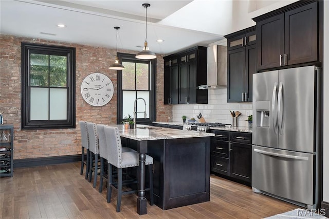 kitchen with stainless steel fridge, wall chimney range hood, a sink, wood finished floors, and light stone counters