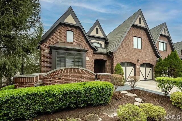 english style home featuring brick siding, a garage, driveway, and roof with shingles