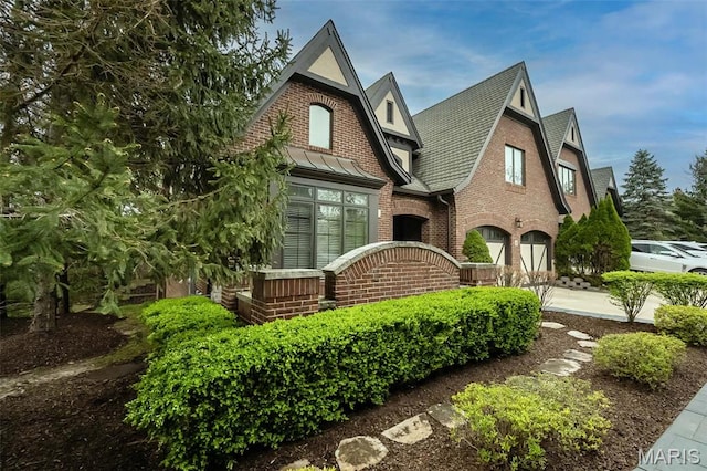 tudor-style house featuring a garage, brick siding, concrete driveway, a standing seam roof, and a shingled roof