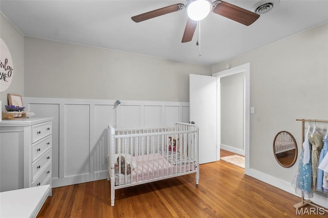 bedroom featuring wood finished floors, a wainscoted wall, a ceiling fan, a crib, and a decorative wall