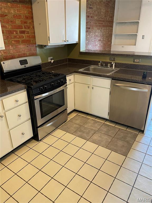 kitchen featuring stainless steel appliances, a sink, open shelves, dark countertops, and brick wall