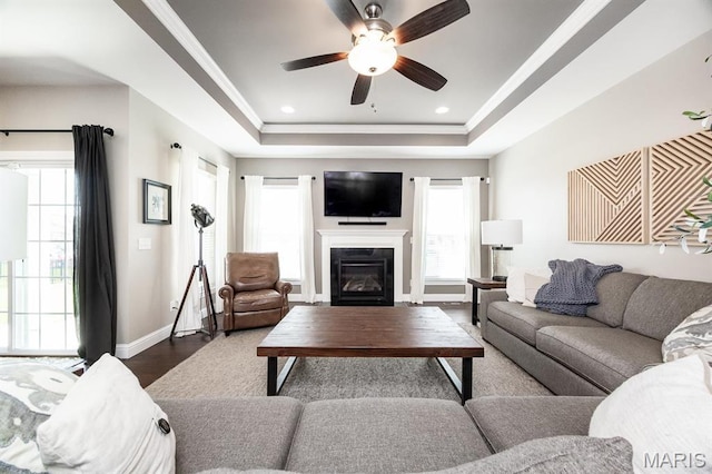 living room with a ceiling fan, a tray ceiling, dark wood finished floors, baseboards, and plenty of natural light