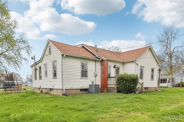 rear view of house featuring a trampoline, cooling unit, a lawn, and a shingled roof