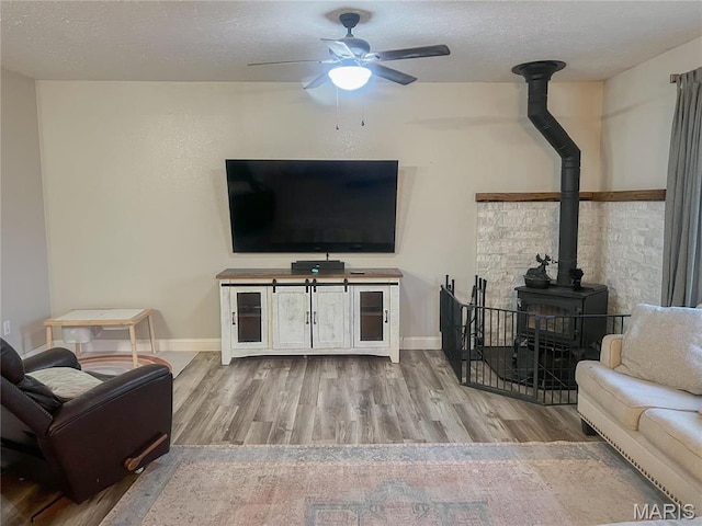 living room featuring a wood stove, wood finished floors, ceiling fan, baseboards, and a textured ceiling