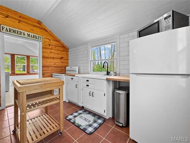 kitchen with white appliances, a sink, lofted ceiling, wood walls, and white cabinetry