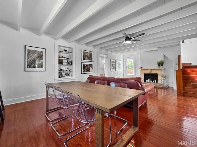 dining area featuring dark wood-type flooring, baseboards, a ceiling fan, a fireplace with raised hearth, and beamed ceiling
