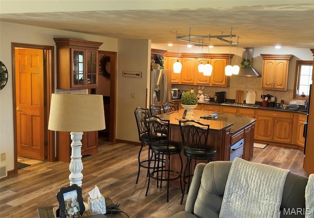 kitchen with wall chimney range hood, a kitchen breakfast bar, backsplash, dark countertops, and wood finished floors