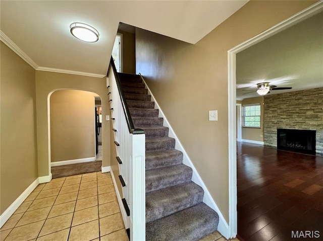 stairs with arched walkways, a ceiling fan, ornamental molding, baseboards, and a stone fireplace
