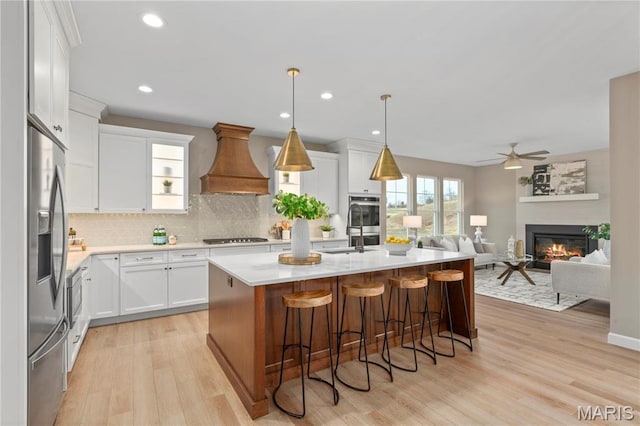 303 Clewer Park Ct, Lake St Louis, MO 63367 - kitchen with custom exhaust hood, stainless steel appliances, a glass covered fireplace, light wood-type flooring, and tasteful backsplash
