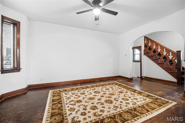 empty room featuring arched walkways, a ceiling fan, baseboards, stairs, and wood finished floors
