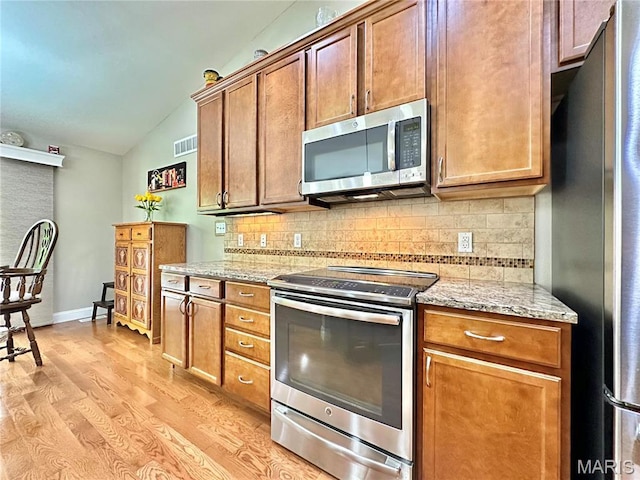 kitchen with appliances with stainless steel finishes, vaulted ceiling, decorative backsplash, light stone countertops, and light wood-type flooring