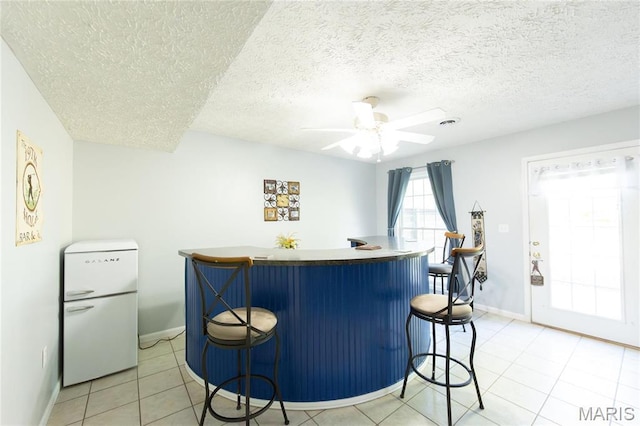 indoor bar with freestanding refrigerator, a ceiling fan, baseboards, light tile patterned floors, and a textured ceiling