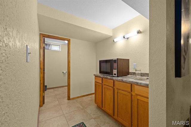 kitchen featuring stainless steel microwave, a textured wall, brown cabinets, light tile patterned floors, and dark countertops