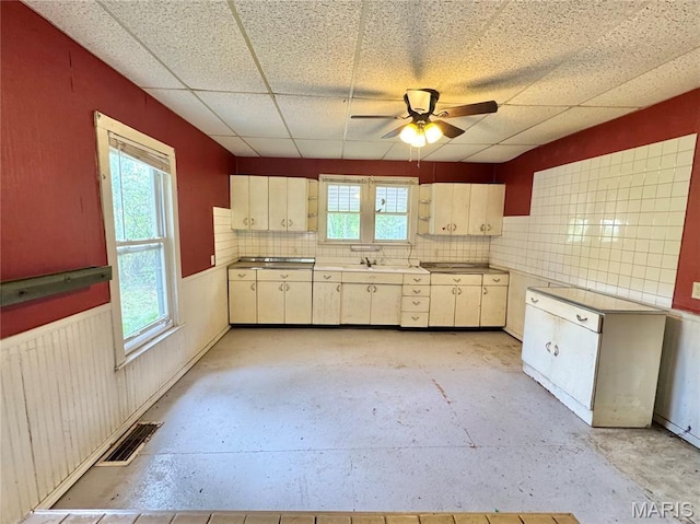 kitchen featuring plenty of natural light, a ceiling fan, a drop ceiling, a wainscoted wall, and tasteful backsplash