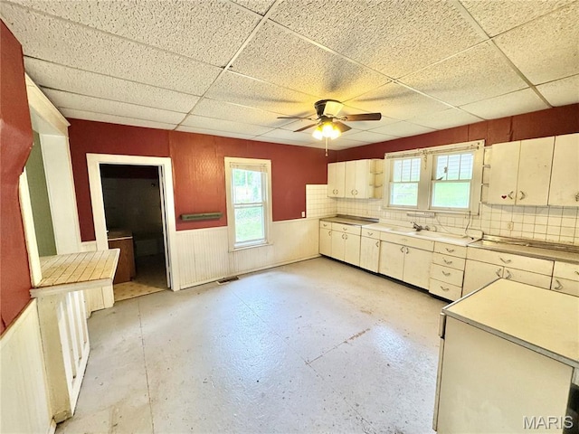 kitchen featuring a paneled ceiling, a ceiling fan, light countertops, a wainscoted wall, and concrete floors