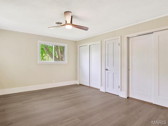 unfurnished bedroom with two closets, wood finished floors, baseboards, crown molding, and a ceiling fan