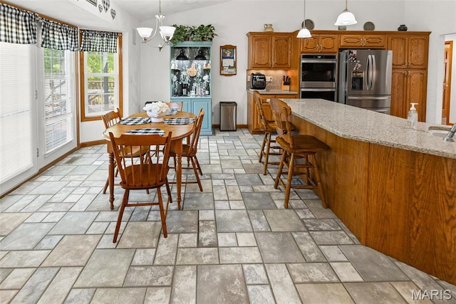 kitchen featuring appliances with stainless steel finishes, stone tile flooring, brown cabinetry, a chandelier, and lofted ceiling