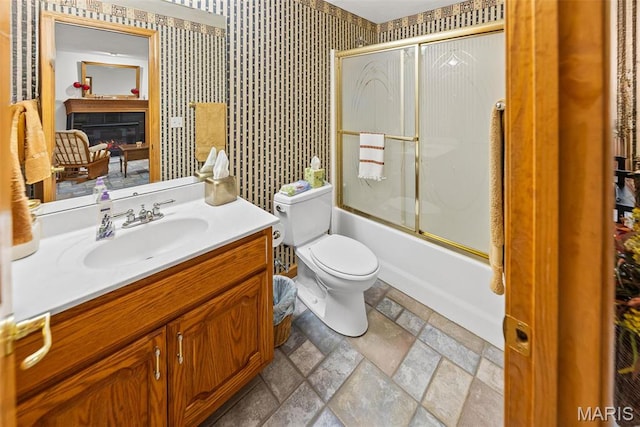 full bath featuring toilet, stone finish floors, vanity, combined bath / shower with glass door, and a glass covered fireplace