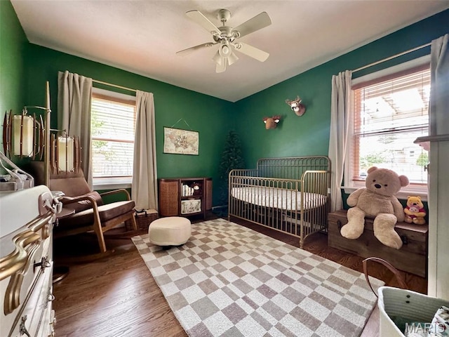 bedroom with multiple windows, wood finished floors, a nursery area, and a ceiling fan