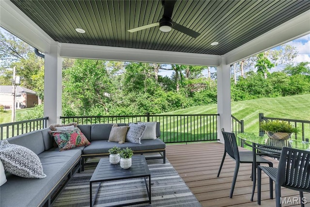 wooden deck with an outdoor living space, a ceiling fan, view of wooded area, and a yard