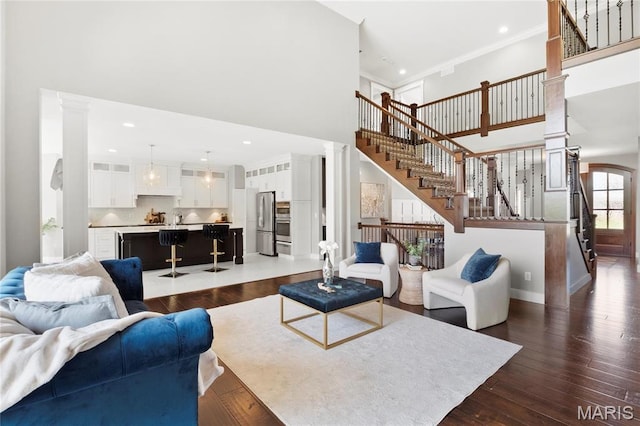 living room featuring a towering ceiling, stairs, decorative columns, dark wood-type flooring, and ornamental molding