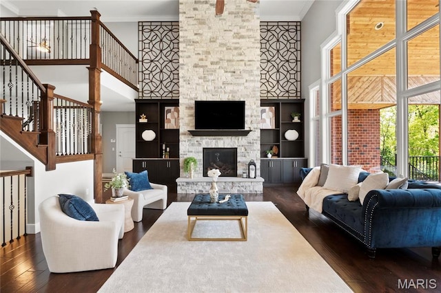 living room featuring dark wood-style floors, a towering ceiling, crown molding, stairs, and a stone fireplace