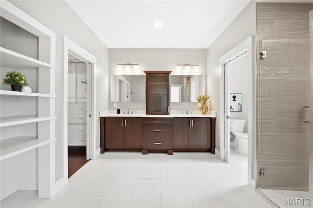 bathroom featuring toilet, double vanity, a tile shower, and baseboards