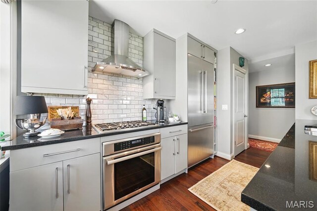 kitchen with stainless steel appliances, wall chimney exhaust hood, decorative backsplash, dark wood-type flooring, and baseboards