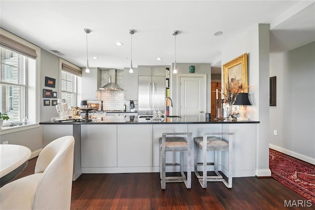 kitchen with wall chimney range hood, built in refrigerator, dark countertops, dark wood-style floors, and a peninsula