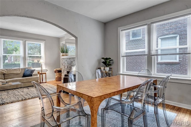 dining room with arched walkways, light wood-type flooring, a stone fireplace, and baseboards