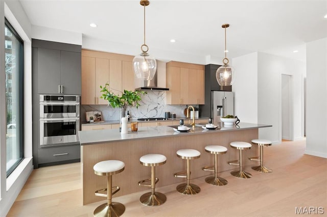215 N Sarah St Unit 12, Saint Louis, MO 63108 - kitchen featuring wall chimney range hood, stainless steel appliances, decorative backsplash, an island with sink, and light wood-style flooring