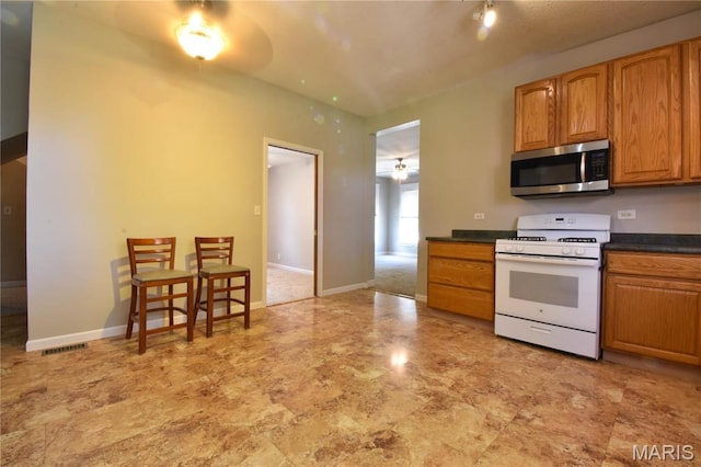 kitchen featuring stainless steel microwave, white gas stove, dark countertops, a ceiling fan, and brown cabinetry