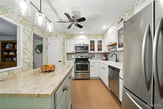 kitchen featuring stainless steel appliances, wallpapered walls, a ceiling fan, a sink, and glass insert cabinets